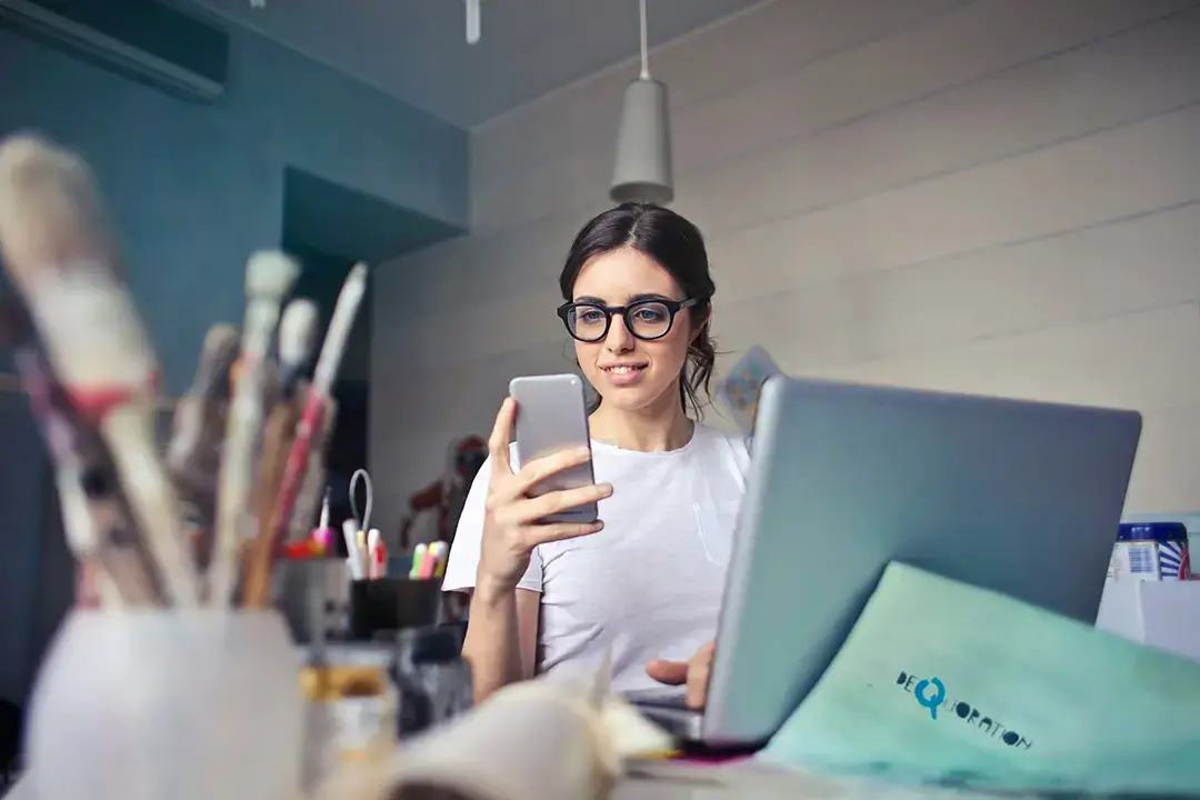 Woman checking phone and laptop at desk.